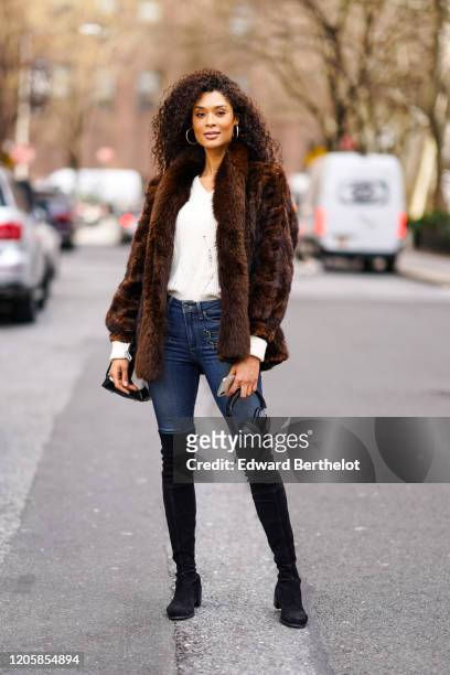 Guest wears a white top, a brown fur coat, blue jeans, black suede thigh high boots, earrings, during New York Fashion Week Fall Winter 2020, on...