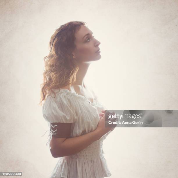 profile view of young woman with long wavy red hair wearing off-shoulder peasant blouse, looking up in hope with hands clasped, studio shot with textured cream backdrop - period costume stock pictures, royalty-free photos & images