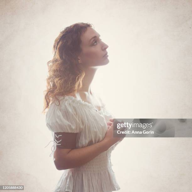 profile view of young woman with long wavy red hair wearing off-shoulder peasant blouse, looking up in hope with hands clasped, studio shot with textured cream backdrop - roupa de época - fotografias e filmes do acervo