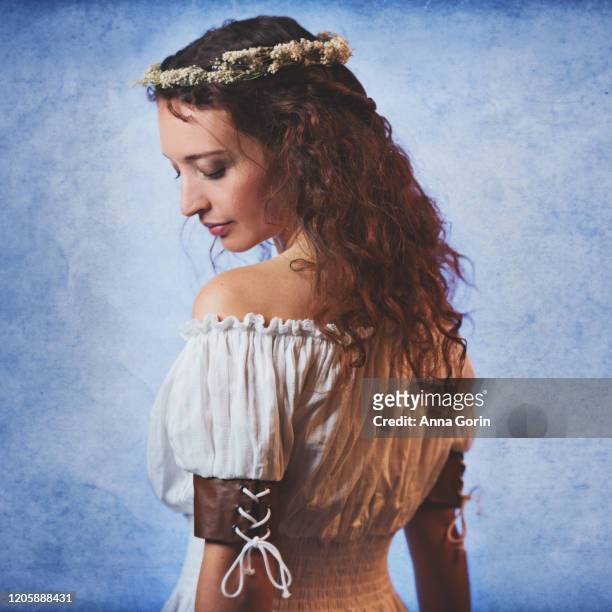 young woman with long wavy red hair wearing off-shoulder peasant blouse and flower crown, looking down at shoulder, studio shot with textured blue backdrop - down blouse ストックフォトと画像