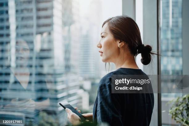 young urban businesswoman using smartphone in the office in front of windows overlooking the city - asian finance stock-fotos und bilder