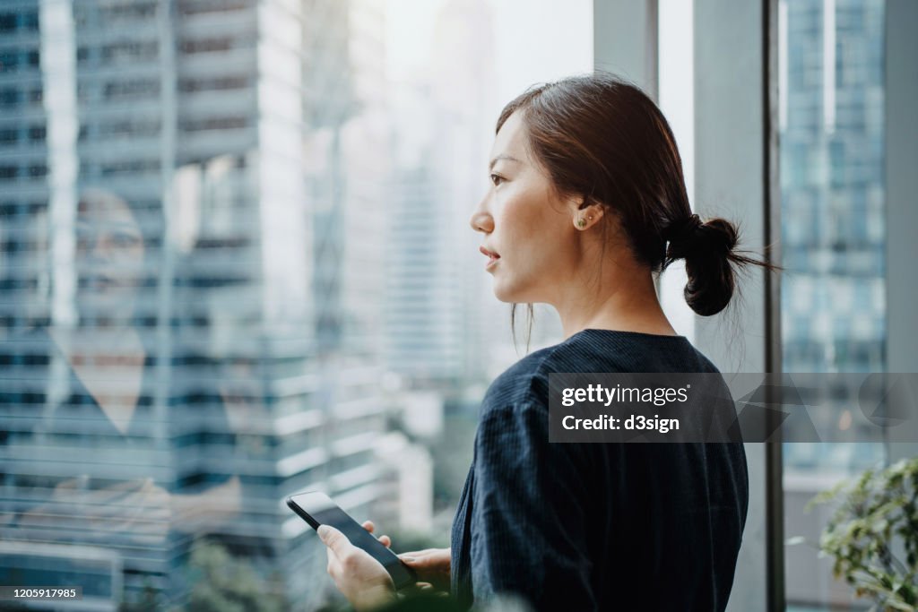 Young urban businesswoman using smartphone in the office in front of windows overlooking the city