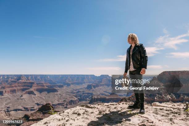 a curly haired blonde man, wearing surprisingly a black leather jacket ,black ripped jeans, black shoes, and a beige linen shirt, is tipping his cowboy hat to the majestic beauty of the grand canyon - cowboy tipping hat stock pictures, royalty-free photos & images