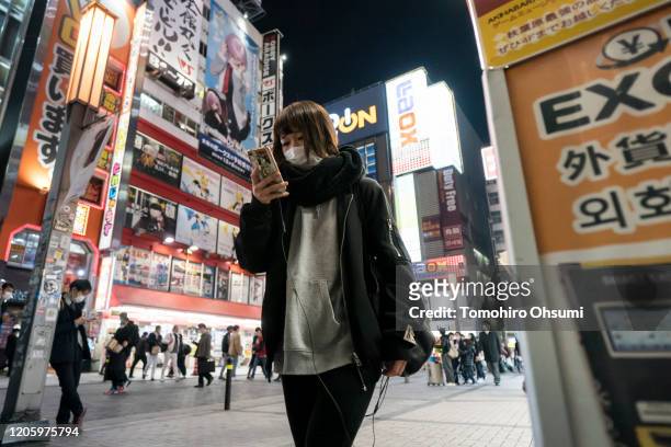 Woman wearing a face mask uses a smartphone as she walks through a shopping district at night on February 13, 2020 in Tokyo, Japan. At least 219...
