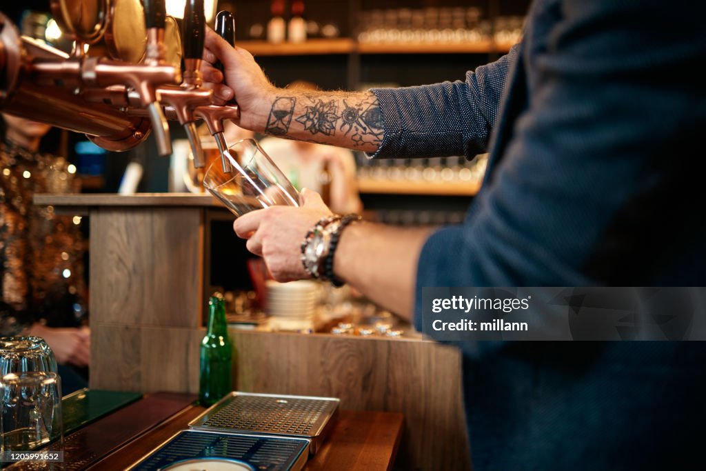 Tattooed caucasian barman pouring beer while standing in pub. Selective focus on hand.