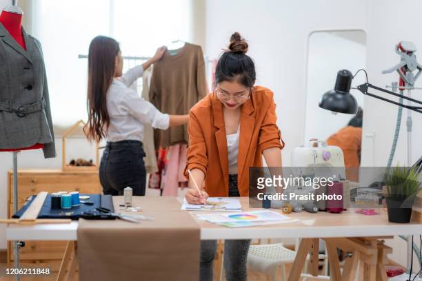 two asian woman at garment factory. they are happy and fashionable. they are standing behind sewing machines and design new dress. small business startup sme entrepreneur or freelance concept - stylist bildbanksfoton och bilder