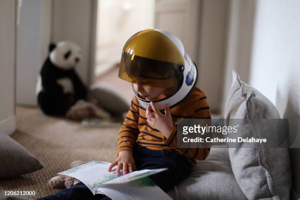 a four year old boy wearing a cosmonaut helmet, reading a book at home - reading fotografías e imágenes de stock