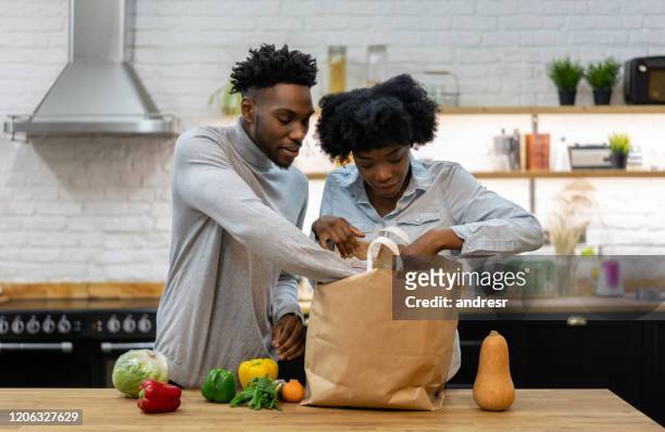 happy african american couple at home unpacking the groceries - young man groceries kitchen stock pictures, royalty-free photos & images
