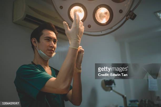 ein asiatischer chinesischer männlicher chirurg, der sich vorbereitet und seine chirurgischen handschuhe im operationssaal anzieht, der durch chirurgisches licht beleuchtet wird - get dressed male stock-fotos und bilder