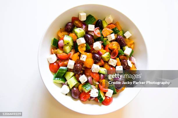 close-up, high-key image of a white bowl filled with a greek salad, including tomatoes, cucumber, olives, onions and feta beans - mediterranean food stock pictures, royalty-free photos & images