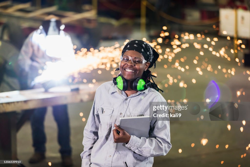 Woman in metal fabrication plant, welder in background