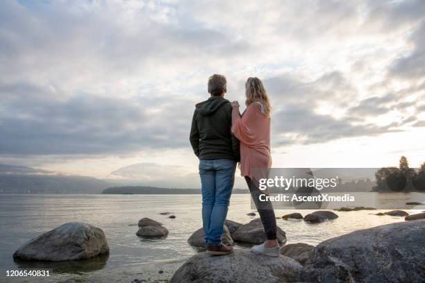 mature couple relax on beach rocks at sunrise - distant stock pictures, royalty-free photos & images