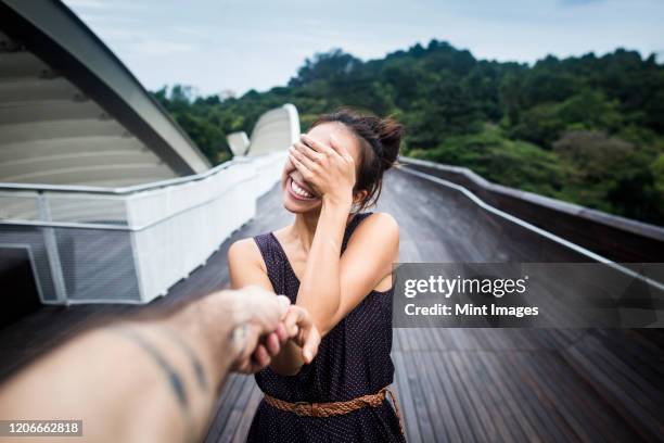 smiling young woman standing on a bridge, covering her face, holding man's hand. - young couple holding hands stock pictures, royalty-free photos & images