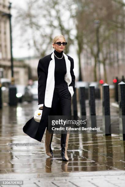 Caroline Daur wears sunglasses, a black turtleneck pullover, a chain metallic necklace, a black long coat with white collar, a white bag, beige and...