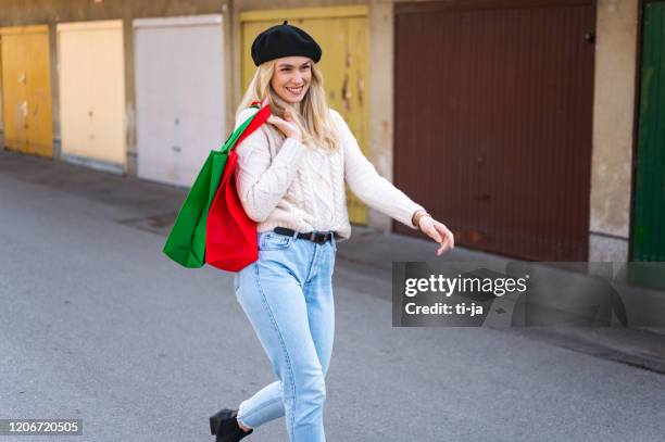 young woman walking with green and red shopping bags outdoors by garages stock photo - beret stock pictures, royalty-free photos & images