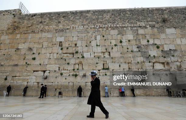 An ultra-Orthodox Jewish man wearing a plstic-covered hat and speaking on a phone walks past people praying at the nearly deserted Western Wall,...