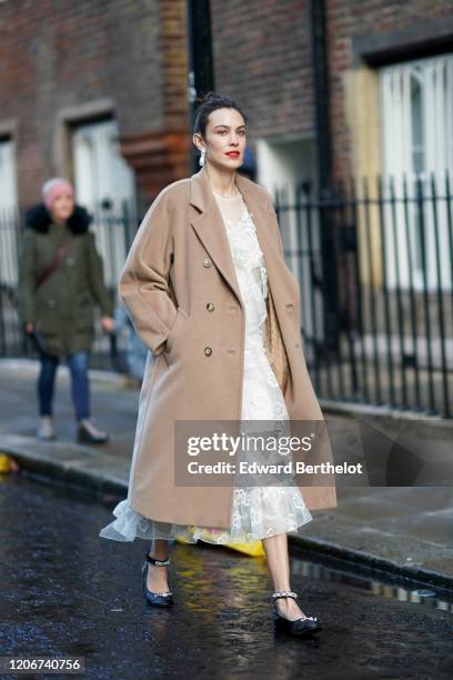 Alexa Chung wears earrings, a pale brown / beige long wool coat, a white lace mesh dress, black bejeweled shoes, during London Fashion Week Fall...