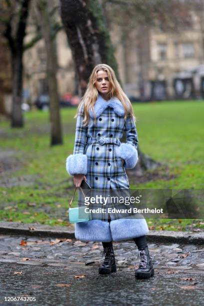 Guest wears a blue checked long coat with fluffy parts, a turquoise bag, a belt, black boots, leggings, during London Fashion Week Fall Winter 2020...