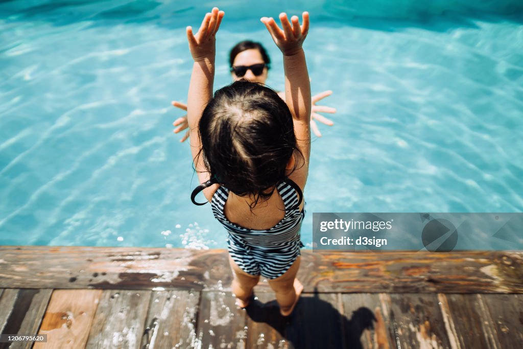 Overhead view of little Asian toddler girl having fun and jumping into her mother's arms in the swimming pool in Summer