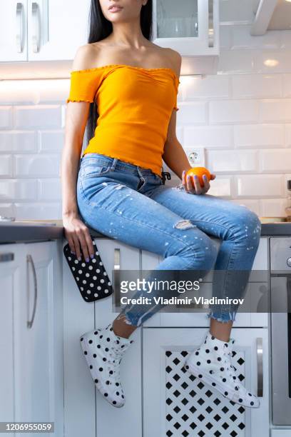 frau mit jeans und turnschuhen mit punkten auf küchentheke sitzend - orange shoe stock-fotos und bilder