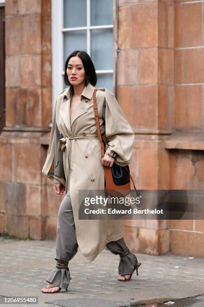 Tiffany Hsu wears a brown bag, a gray and beige trench coat, gray pants, shoes, outside GW Anderson, during London Fashion Week February 2020 on...