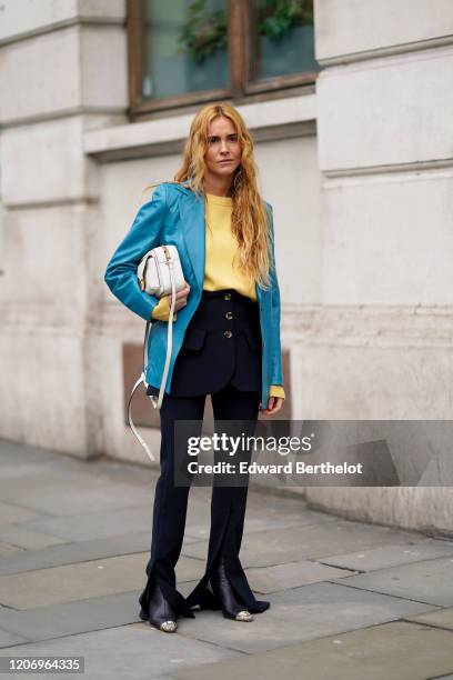 Blanca Miro wears a blue blazer jacket, a yellow pullover, a dark short skirt with buttons, dark flared pants, pointy boots, a white bag, during...