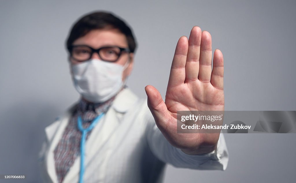 An Asian Or Chinese Doctor In A Medical Mask And Glasses Shows A Warning Stop Sign With His Hand.