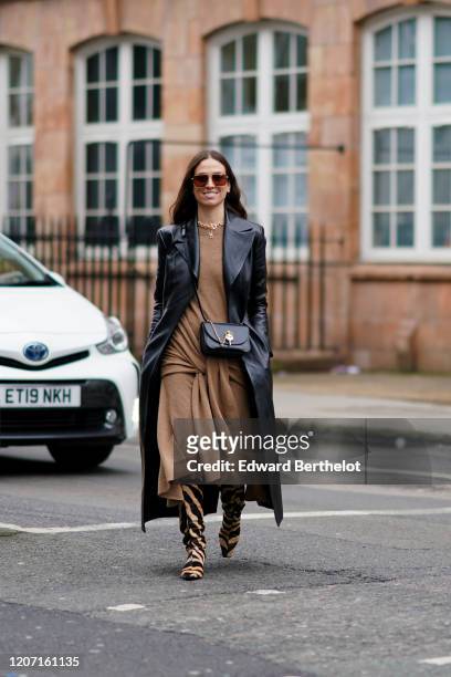 Erika Boldrin wears sunglasses, a brown wool long dress, a black leather long coat, a bag, zebra print brown and black striped boots, during London...