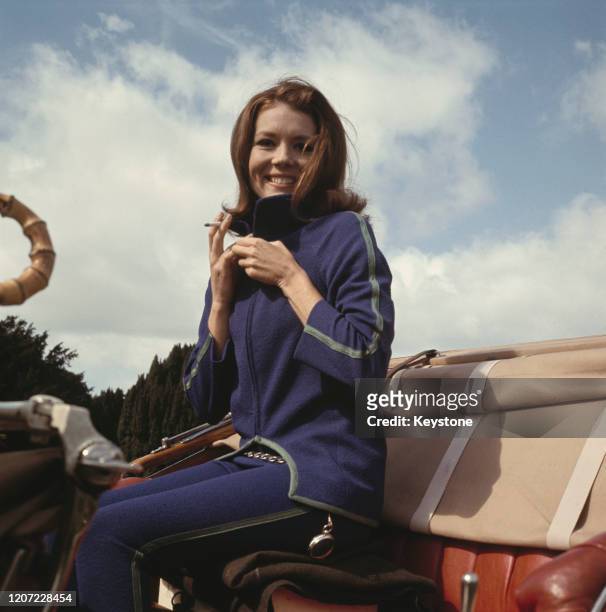 British actress Diana Rigg wearing a blue tracksuit with green trim, holding a cigarette while seating in the driver's seat of a car during filming...