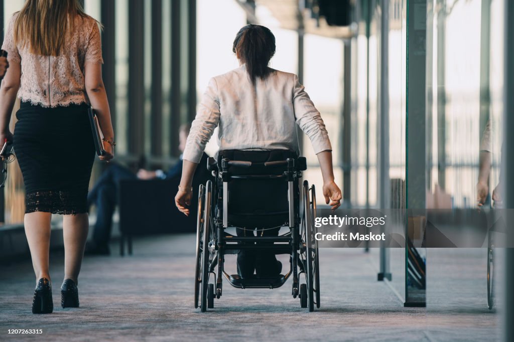Rear view of female entrepreneur walking with disabled businesswoman in corridor at workplace