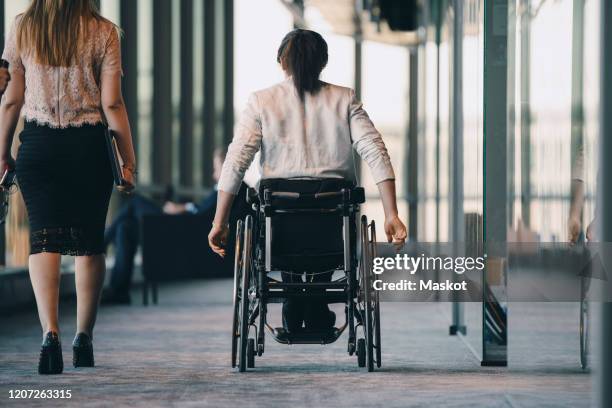 rear view of female entrepreneur walking with disabled businesswoman in corridor at workplace - körperliche behinderung stock-fotos und bilder