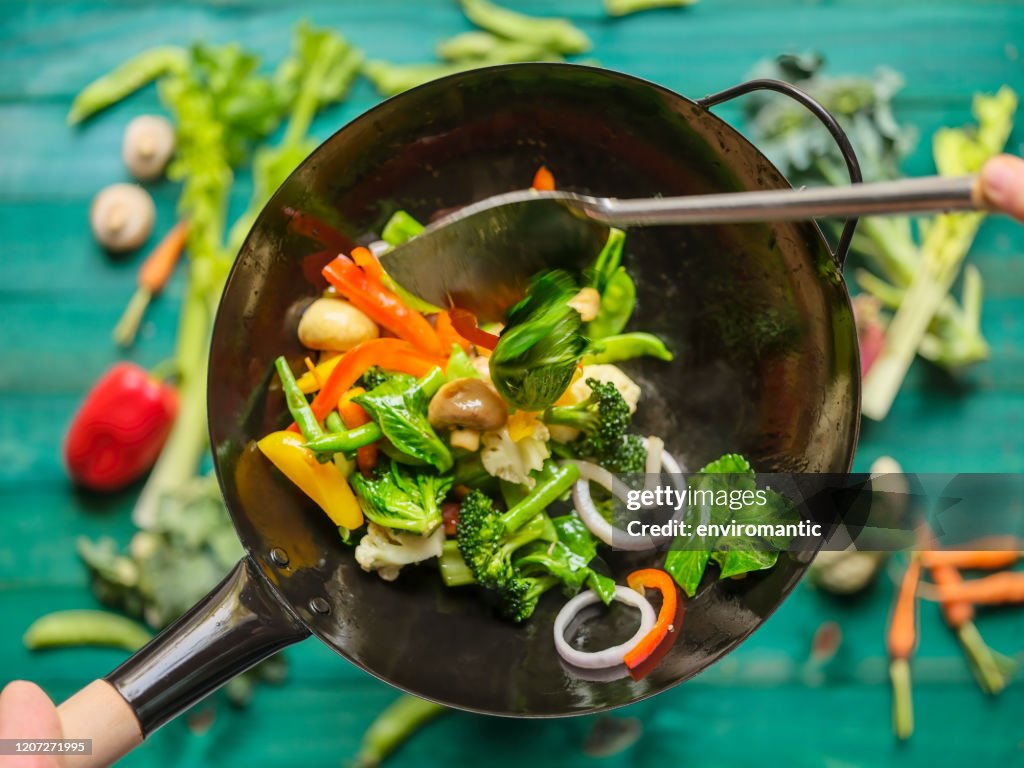 Rühren Braten und sauteing eine Vielzahl von frischen bunten Markt Gemüse in einem heißen dampfenden Wok mit Gemüse auf einem türkisfarbenen Holztisch Hintergrund unter dem Wok.
