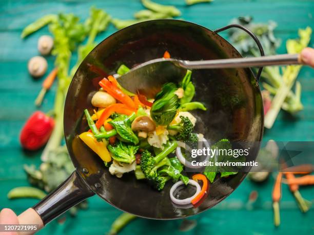 roer bakken en sauteing een verscheidenheid van verse kleurrijke marktgroenten in een hete stomende wok met groenten op een turquoise gekleurde houten lijstachtergrond onder de wok. - wok stockfoto's en -beelden