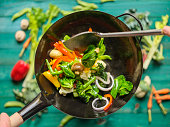Stir frying and sauteing a variety of fresh colorful market vegetables in a hot steaming wok with vegetables on on a turquoise colored wood table background below the wok.