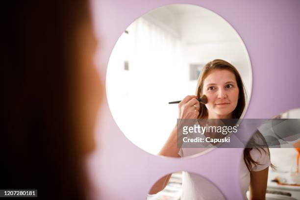 jonge vrouw die blush draagt die in de spiegel bij slaapkamer kijkt - maquiagem stockfoto's en -beelden