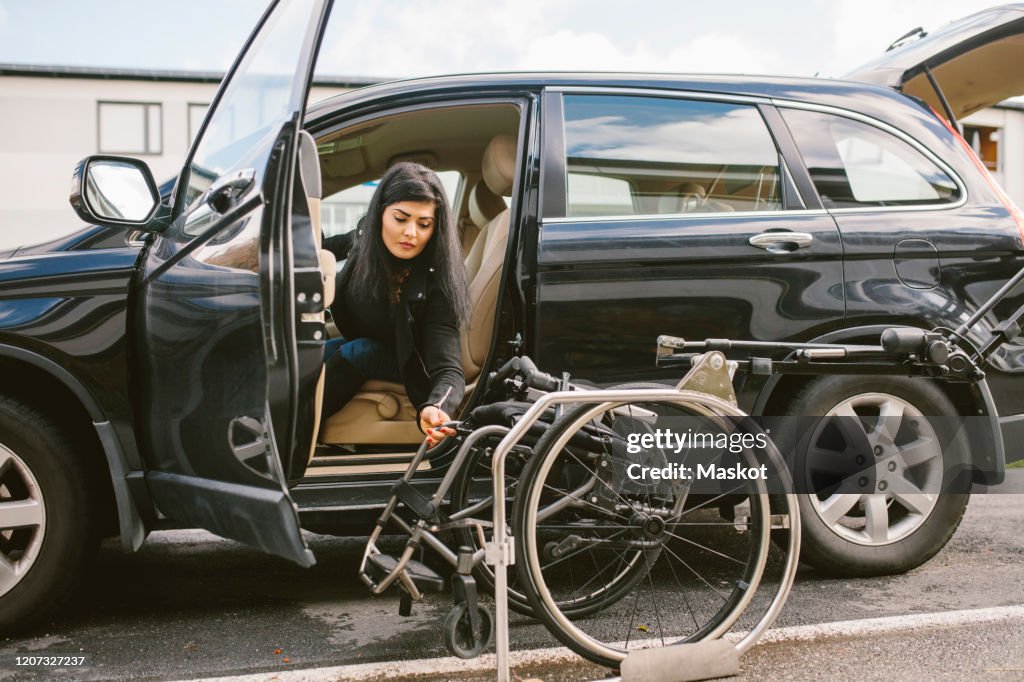 Young disabled woman holding wheelchair by car on roadside in city