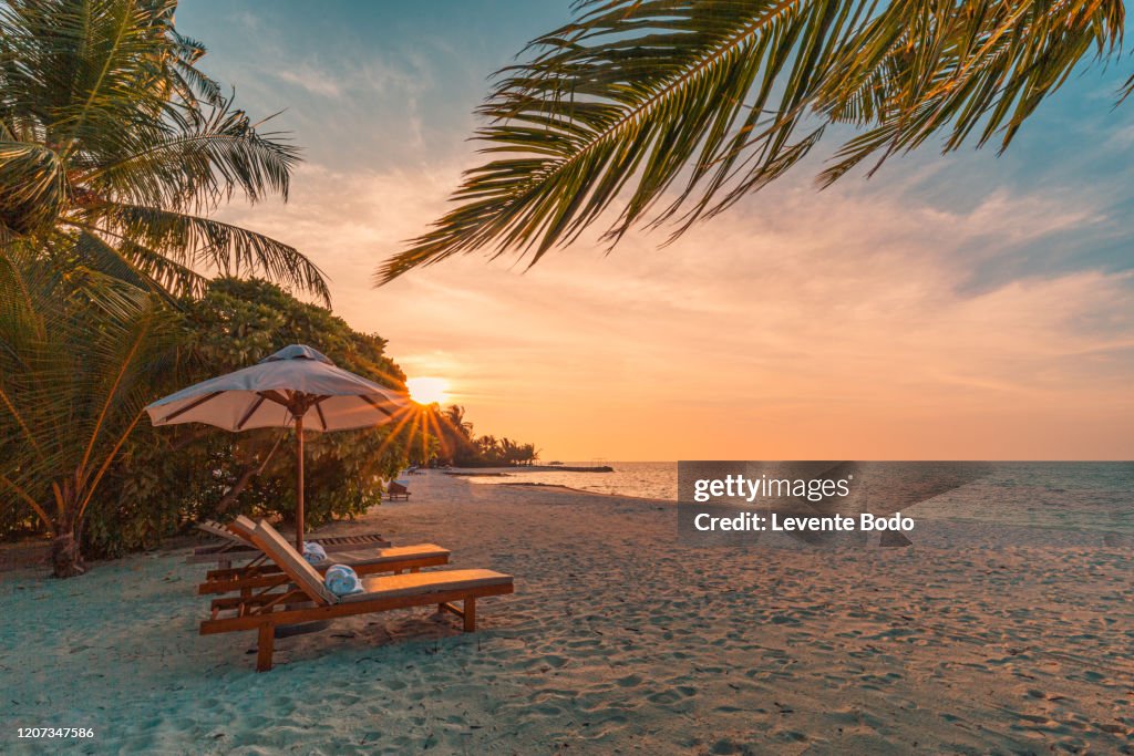Beautiful tropical sunset scenery, two sun beds, loungers, umbrella under palm tree. White sand, sea view with horizon, colorful twilight sky, calmness and relaxation. Inspirational beach resort hotel