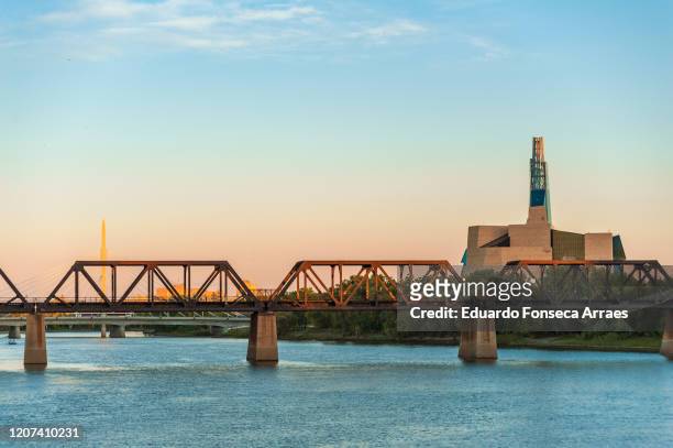 view of the red river, a railway bridge, the esplanade riel bridge on the background and the canadian museum for human rights on the right - esplanade riel fotografías e imágenes de stock