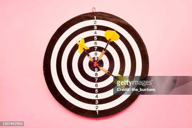 high angle view of a dartboard and three yellow darts on pink colored background - ziel stock-fotos und bilder