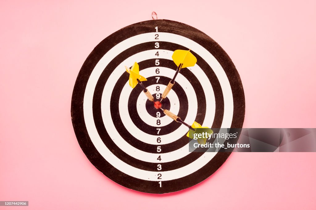 High angle view of a dartboard and three yellow darts on pink colored background
