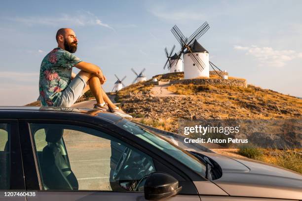 bearded man sits on the roof of the car on the background of windmills in consuegra, mancha, spain - don quijote de la mancha fotografías e imágenes de stock