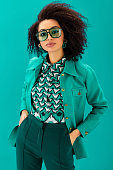 african american woman in jacket with hands in pockets looking at camera isolated on turquoise