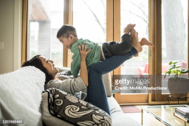 mother playing with young son (2 yrs) on couch at home - sollevare foto e immagini stock