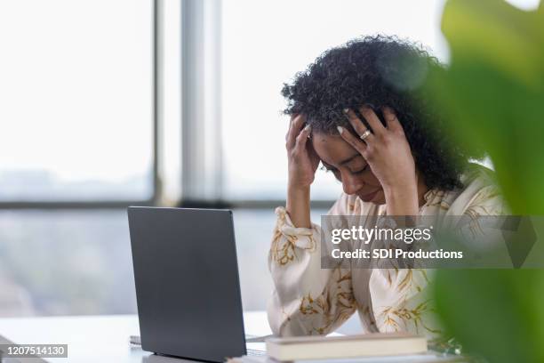 mature woman holds head in hands while sitting at desk - black women praying stock pictures, royalty-free photos & images