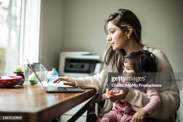 mother multi-tasking with infant daughter in home office - madre ama de casa fotografías e imágenes de stock