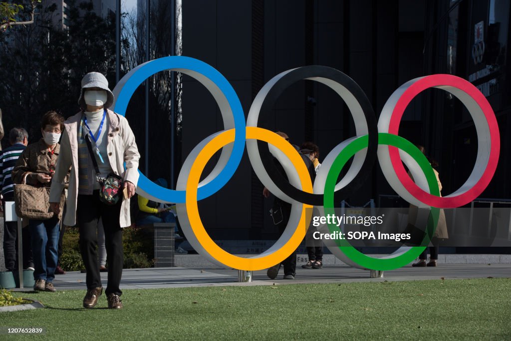 Women with surgical masks walk next to the Olympic Rings in...