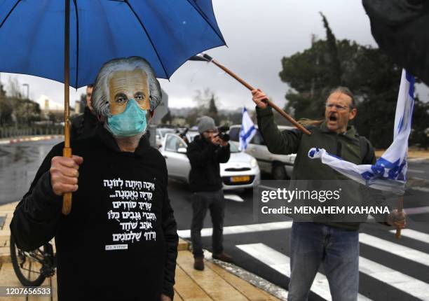 Man holding an umbrella, wearing a protective face mask beneath another mask showing the face of late physicist Albert Einstein, and a hoodie sweater...