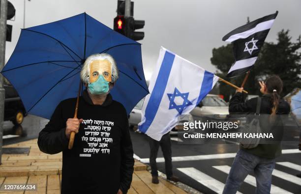 Man holding an umbrella, wearing a protective face mask beneath another mask showing the face of late physicist Albert Einstein, and a hoodie sweater...