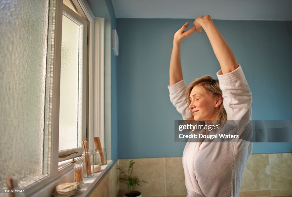 Woman stretching arms with eyes closed in morning bathroom.