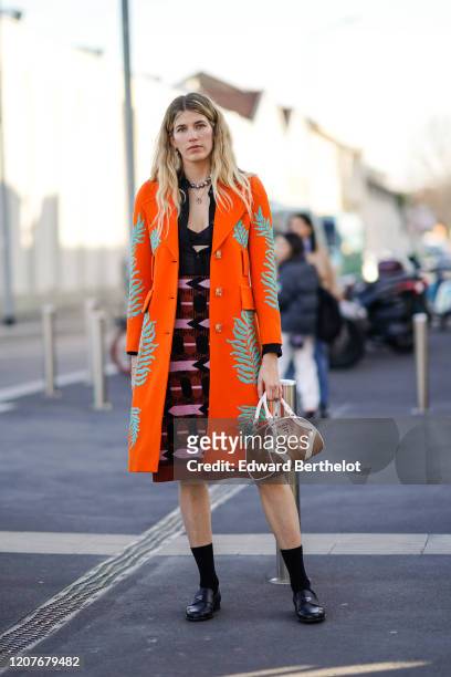 Veronika Heilbrunner wears an orange long coat, a pink and purple dress, a brown leather Prada bag, black socks, black leather shoes, outside Prada,...